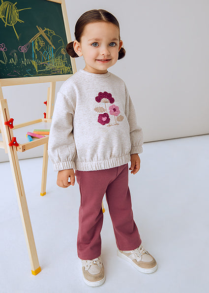 Child wearing a floral sweater and maroon pants standing in front of a blackboard with drawings.